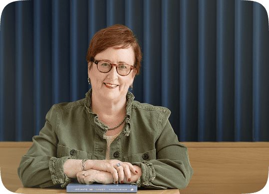 Kathleen Quinn seated at a table with hands folded and smiling, wearing glasses and a green jacket, conveying professionalism, warmth, and trust.