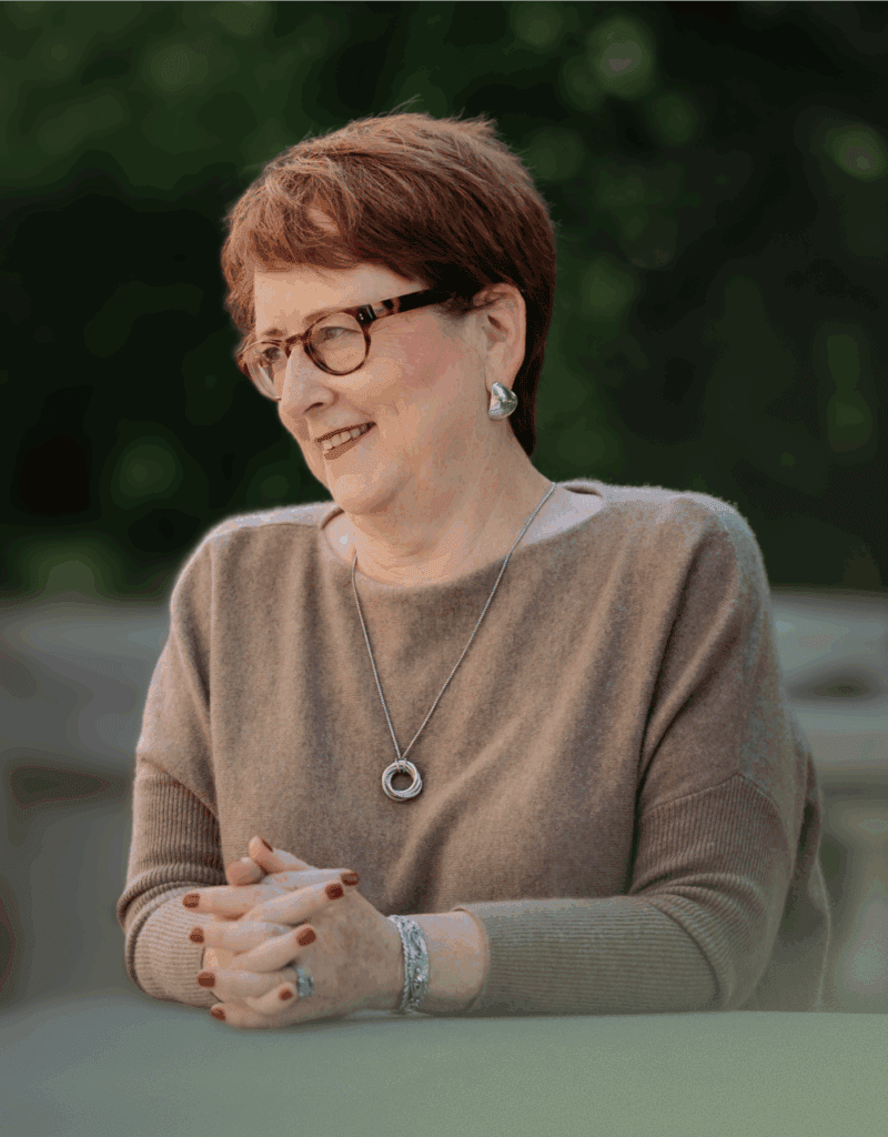 Professional portrait of a woman sitting outdoors, smiling with hands folded on a table.