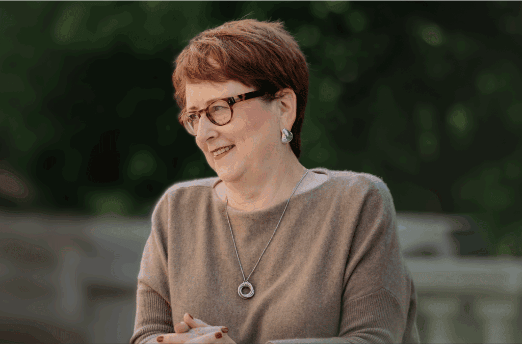 Professional portrait of a woman sitting outdoors, smiling with hands folded on a table.