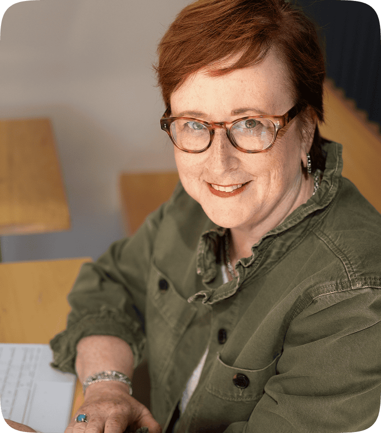 Kathleen Quinn smiling while seated at a desk, wearing glasses and a green jacket, representing approachability, focus, and leadership in her coaching work.