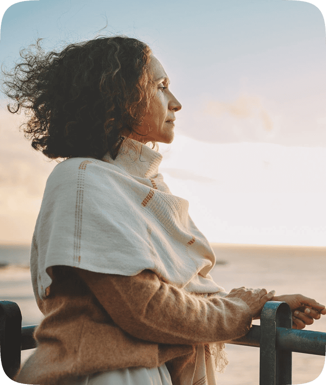 A thoughtful middle-aged woman stands by a seaside railing at sunset, wrapped in a beige shawl and sweater, looking toward the horizon — symbolizing reflection, transition, and personal growth.
