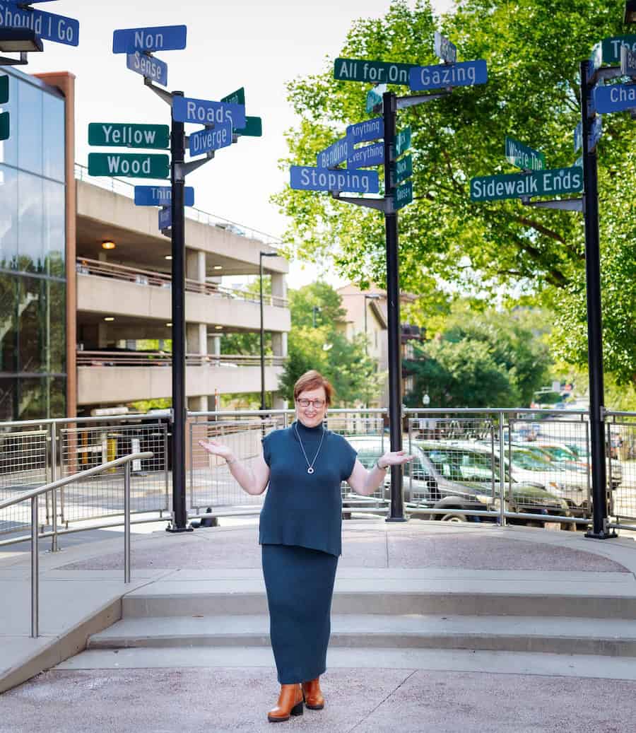Kathleen Quinn standing outdoors beneath multiple street signs, smiling with open arms, symbolizing choice, transition, and personal direction in leadership and life.