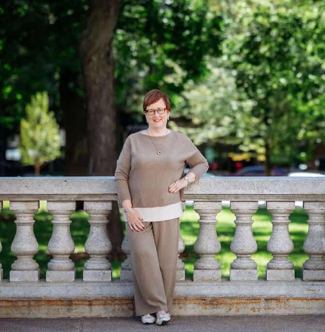 Kathleen Quinn standing by a stone railing in a park, smiling warmly in neutral attire, representing confidence, calm, and authenticity.
