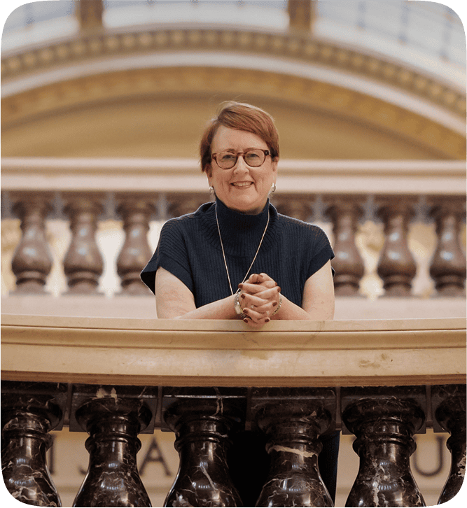 Kathleen Quinn standing confidently on a balcony with elegant architecture, smiling warmly and conveying professionalism and leadership presence.